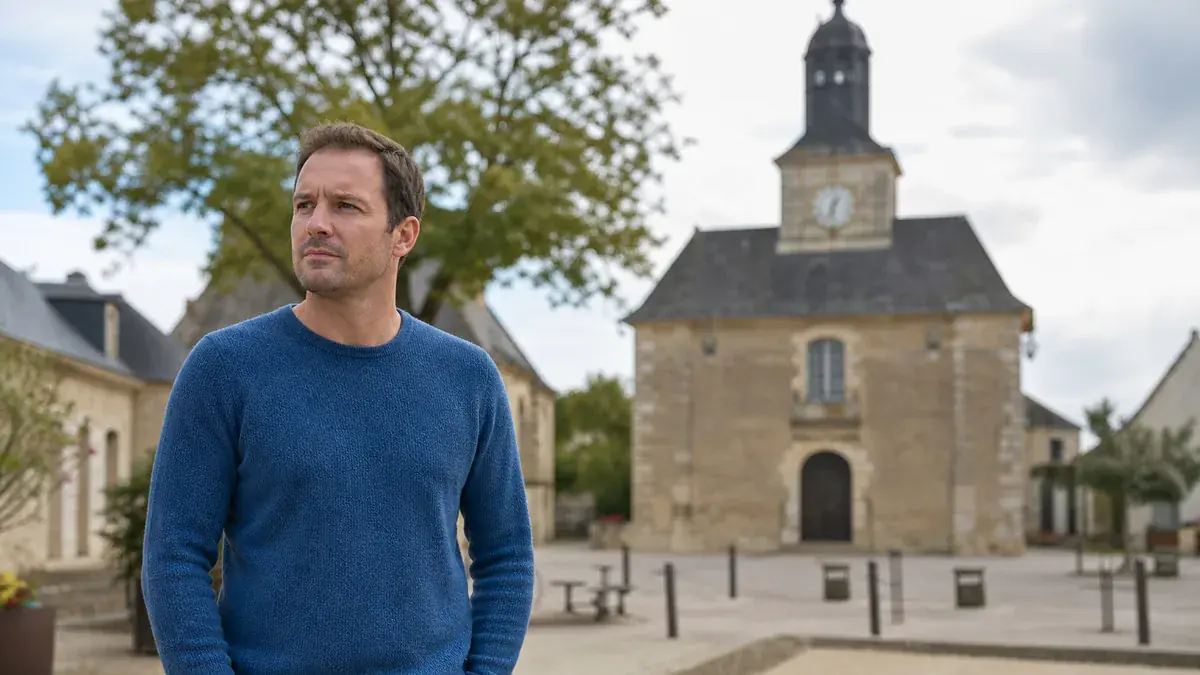 Homme en pull bleu debout sur une place de village, regard sérieux tourné sur le côté, église en arrière-plan