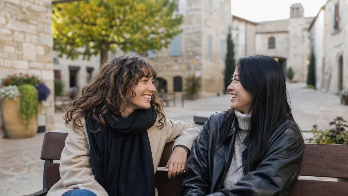 Deux amies assises sur un banc dans une ruelle en pierre, échangeant un regard complice et souriant
